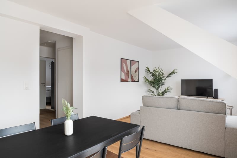 Spacious living room with white walls, hardwood floors, and a large window. The room features a gray sofa, a black dining table, and a TV stand with a television. There are also some potted plants and framed artwork on the walls.