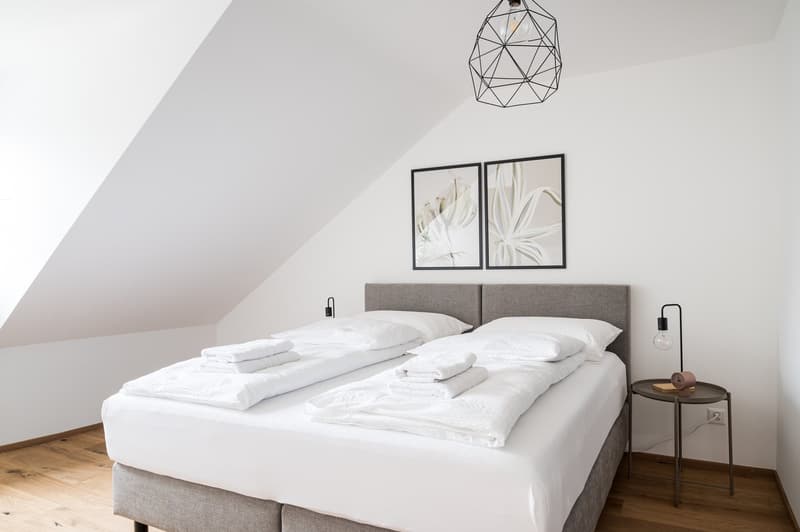 Spacious bedroom with white bedding, gray upholstered headboard, framed artwork on the wall, and a geometric pendant light fixture.