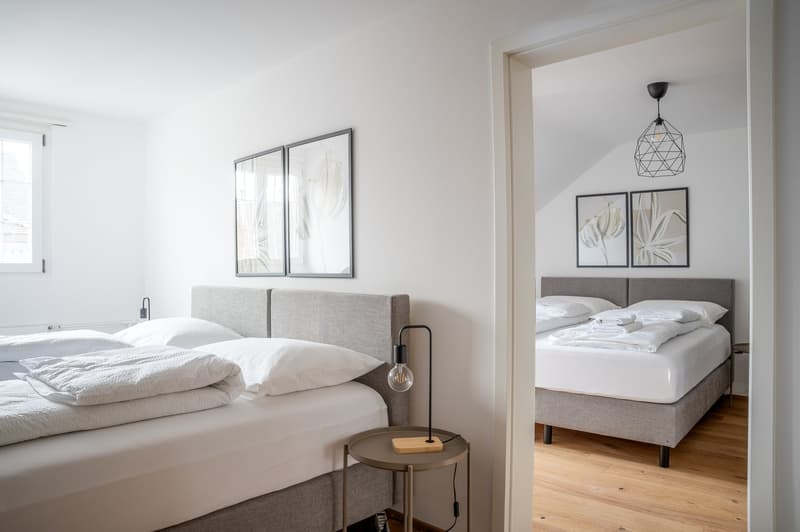 Spacious bedroom with white walls, hardwood floors, large windows, gray upholstered bed frame, side tables, and modern pendant lighting. The room also features framed artwork on the walls and an open doorway leading to an adjacent bedroom.