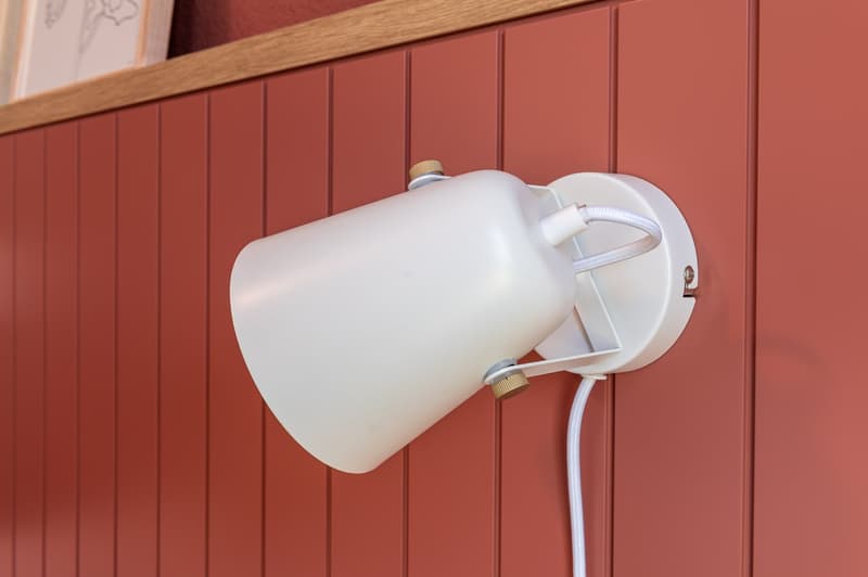 A white wall lamp with adjustable head is mounted on a red wall panel. The lamp has a cylindrical base and a cylindrical reflector, both of which are white. The cord of the lamp is white.