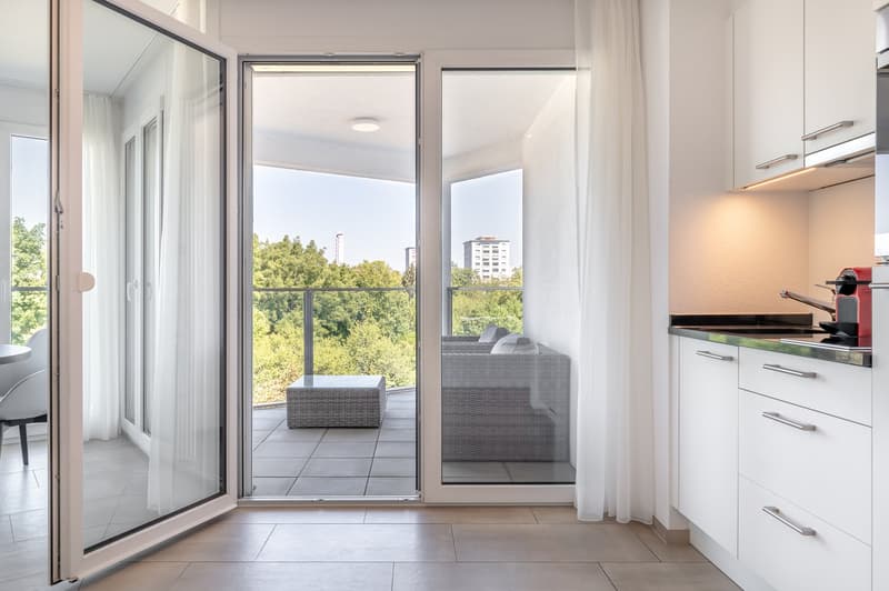 open door to balcony, gray sofa, tile floor, trees and buildings outside, large windows