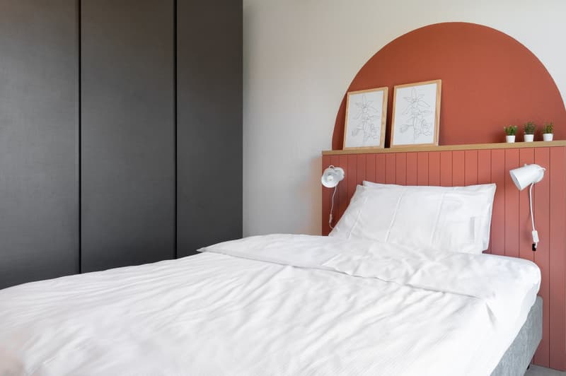 minimalist bedroom with white bedding, dark grey wardrobe, pink headboard, wall lamps, framed art, and potted plants