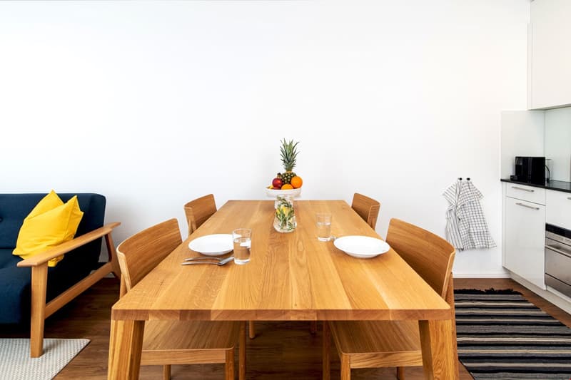 light wooden table, chairs, white walls, open to living area, bowl of fruit, glass of water, plate