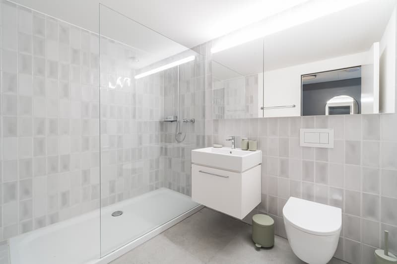 white tiled walls, glass shower enclosure, white sink, white toilet, green footstool