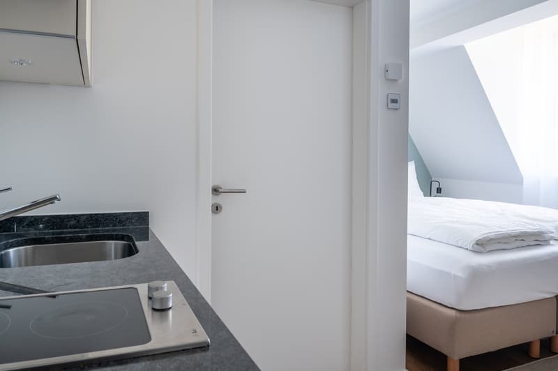 kitchen area with sink, induction hob, white walls, white door, bed with white bedding