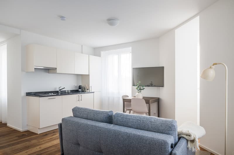 modern kitchen with white cabinets, wooden floor, gray couch, dining table, TV, lamp