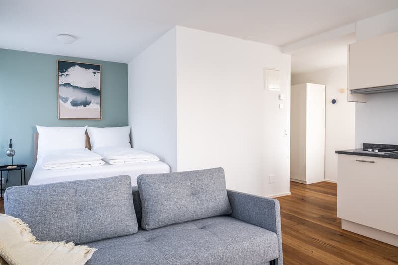 A small bedroom with a double bed, a gray sofa, a nightstand, a picture frame, and a kitchen area with a countertop and cabinets.