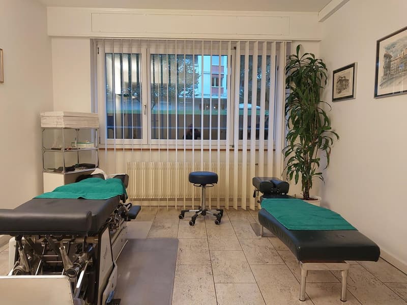 Medical examination room with examination tables, medical equipment, adjustable stool, large window with blinds, and decorative plant.