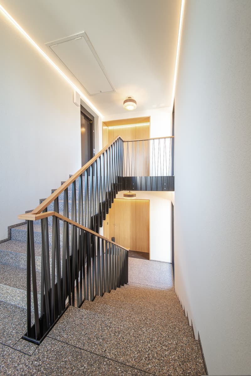 modern staircase with wooden handrail and black metal railings, granite steps, white walls, ceiling lights