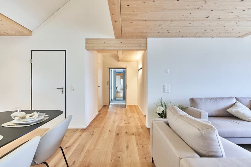 Open-concept living space with hardwood floors, white walls, and exposed wooden beams on the ceiling. The room features a dining area with a black table and chairs, and a living area with a comfortable sofa. There is a doorway leading to a balcony or terr