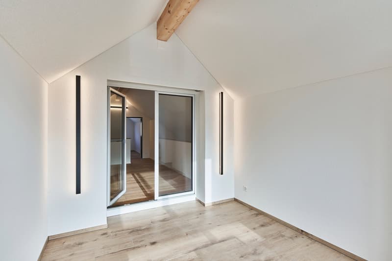 Spacious bedroom with slanted ceiling, wooden beams, and large sliding glass doors leading to a balcony or terrace. The room has a light and airy feel with hardwood flooring and white walls.