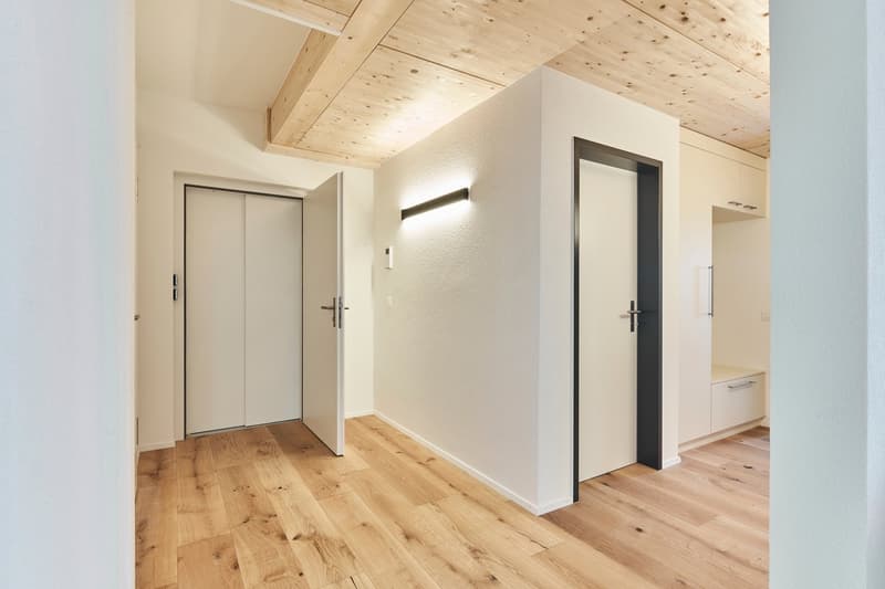 The image shows a hallway with a wooden ceiling, hardwood floors, and white walls. There are two doors visible, one on the left and one on the right. The hallway appears to be part of a larger living space.
