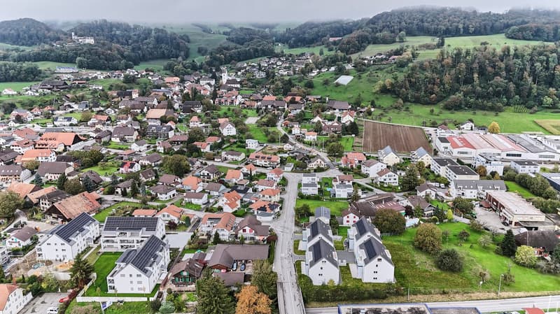 This image shows an aerial view of a small town nestled in a hilly, forested landscape. The town consists of a dense cluster of residential and commercial buildings with red-tiled roofs, surrounded by green fields and forests. There are several large indu