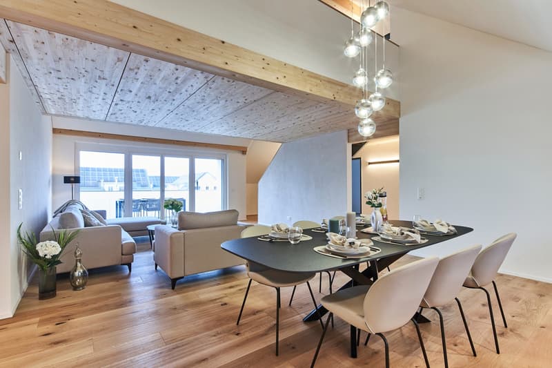 Spacious open-plan living and dining area with wooden beamed ceiling, large windows, hardwood floors, modern furniture including a sofa and dining table with chairs, and a chandelier lighting fixture.
