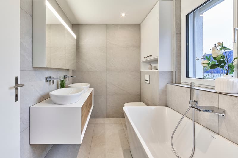 Spacious bathroom with modern design, large vanity with double sinks, freestanding bathtub, tiled walls, and large window providing natural light