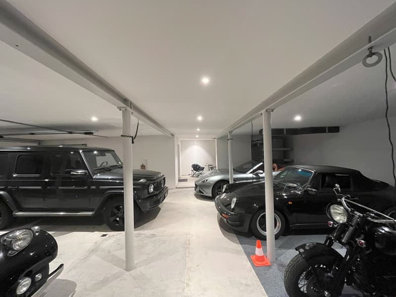 Underground parking garage with four parking spaces, two motorcycles parked, white painted walls, concrete floor, pillars, recessed ceiling lighting
