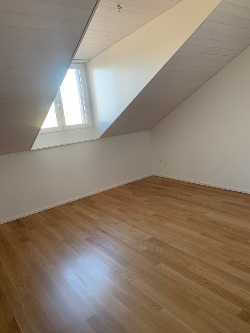 The image shows a bedroom with a slanted ceiling and a large window. The room has hardwood flooring and white walls. The space appears to be clean and well-lit.