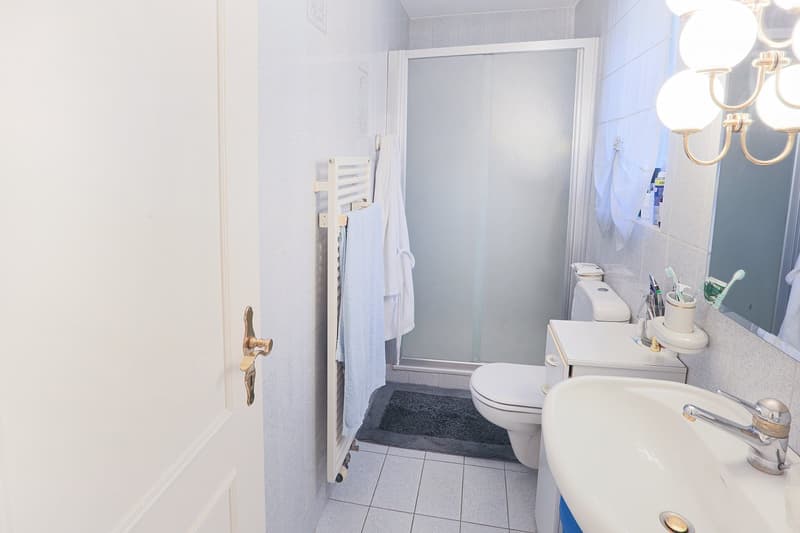 Bathroom with white tiles, white sink, white toilet, glass shower, towel rack, mirror, chandelier