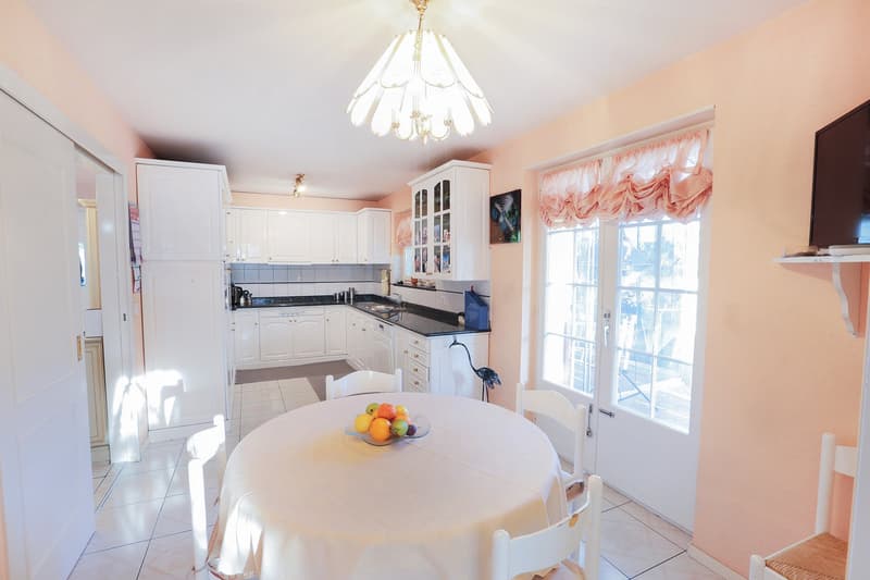 white kitchen with white cabinets, black countertops, white dining table, white chairs, chandelier, pink walls, white floor, window with curtains