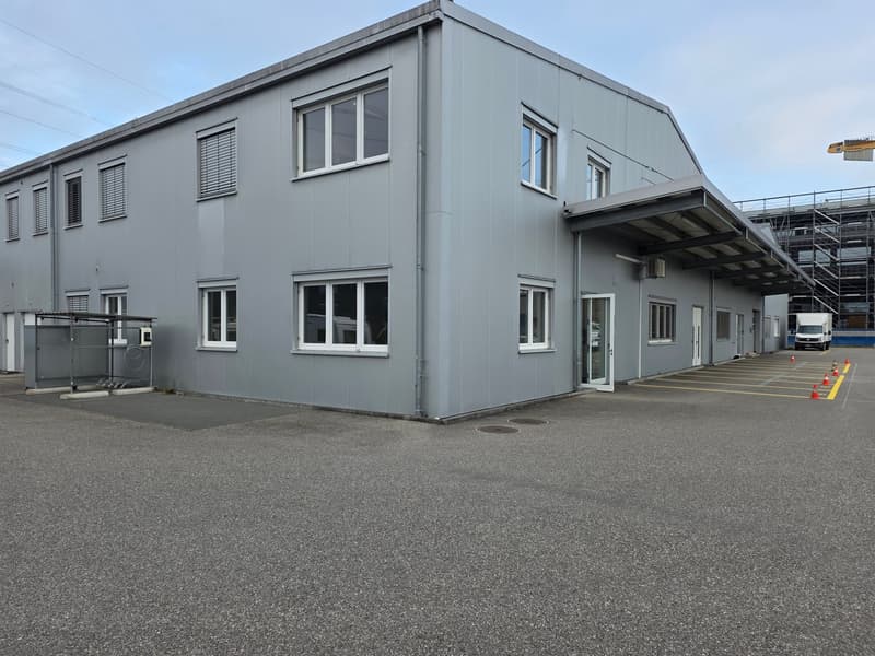 large gray industrial building, with several windows, entrance, and parking lot in front
