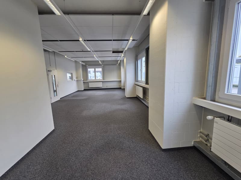 An empty room with gray carpeting, white walls, and ceiling lights. There are windows and radiators along the wall, and a door on the left side.
