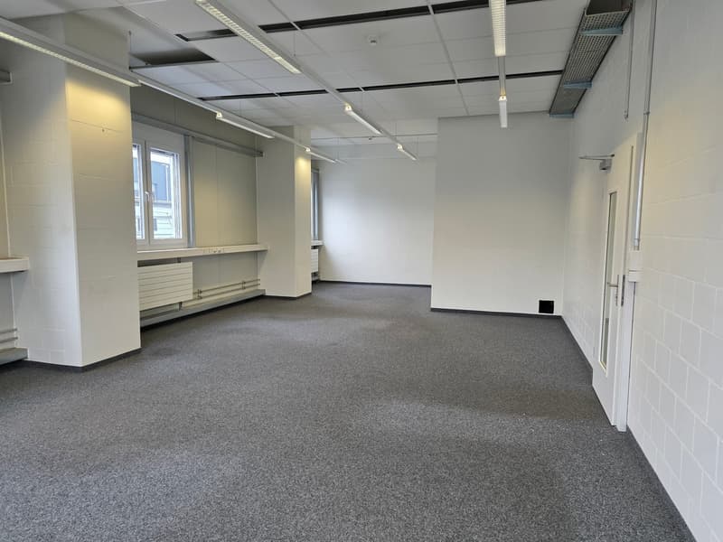 Empty room with gray carpeted floor, white walls, florescent ceiling lights, windows, and an empty desk.
