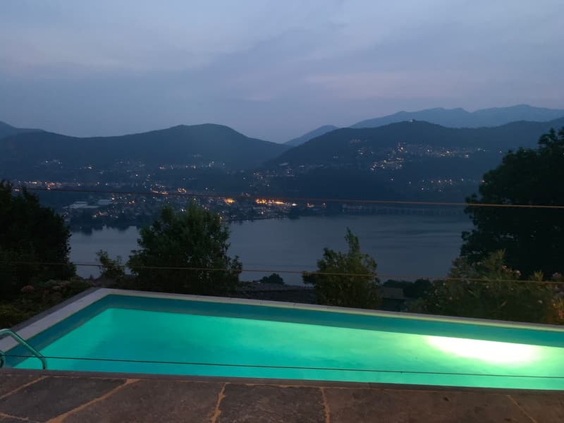 The image shows a swimming pool with a stunning view of a mountainous landscape in the background. The pool is illuminated, creating a serene and tranquil atmosphere. The surrounding area appears to be a residential property with lush greenery and a view