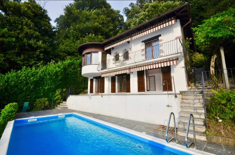 Two-story house with a balcony, surrounded by lush greenery. The house has a swimming pool in the front yard.
