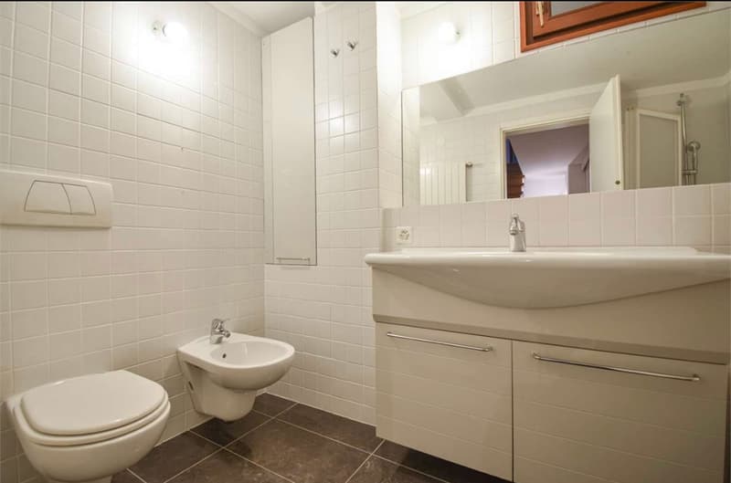 The image shows a modern bathroom with white tile walls, a vanity with a sink, a toilet, and a mirror above the vanity.
