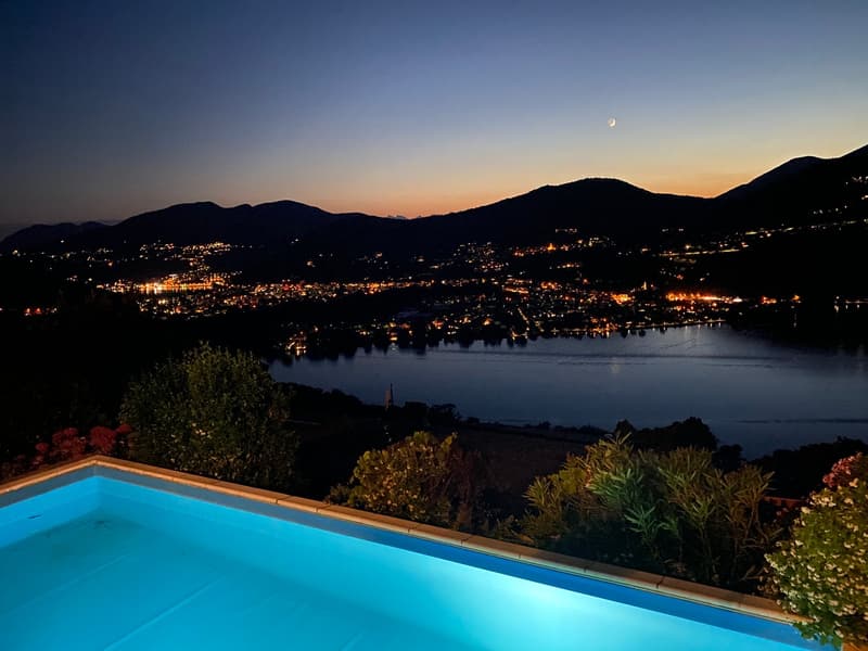 Luxury villa with an infinity pool overlooking a scenic lake surrounded by mountains at night. The pool is illuminated, and the town below is lit up with twinkling lights. A crescent moon is visible in the sky.