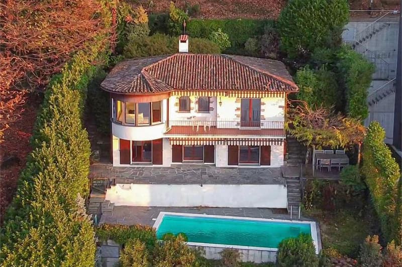 Two-story house with stone exterior, red tile roof, large windows, and a swimming pool in the backyard surrounded by lush greenery and landscaping.