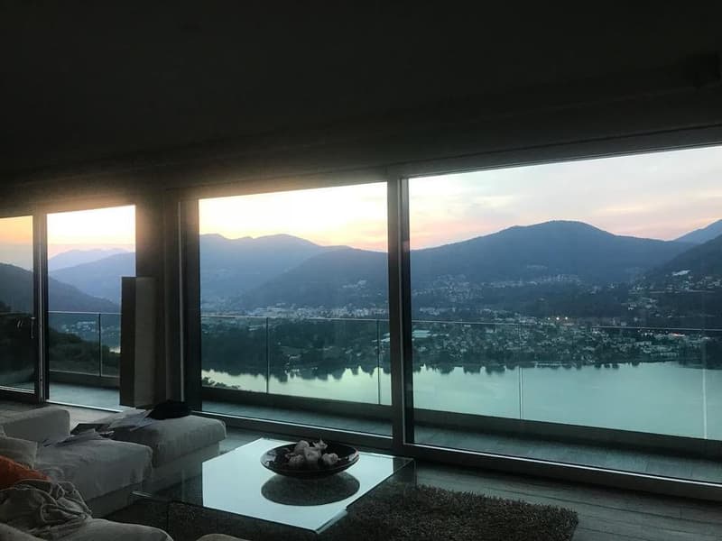 Large windows with a panoramic view of a lake surrounded by mountains, cozy living room with a glass coffee table and a bowl of decorative objects