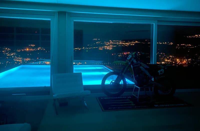 Large windows overlooking a night view of a city, with a motorcycle parked in the foreground and a swimming pool visible outside