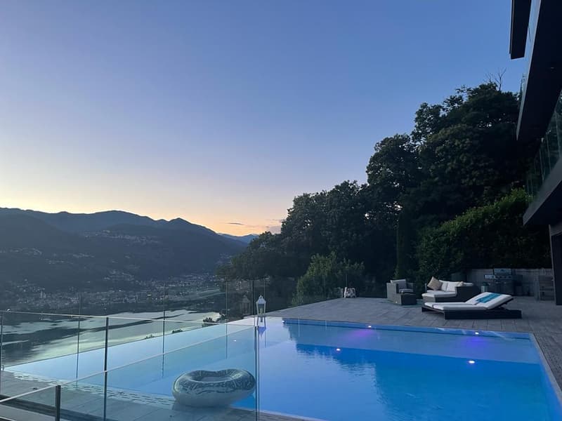The image shows a luxurious outdoor pool area with a stunning view of the surrounding mountains and landscape. The pool is surrounded by a glass railing and has a deck with lounge chairs. The sky is clear and blue, indicating a beautiful sunset or sunrise
