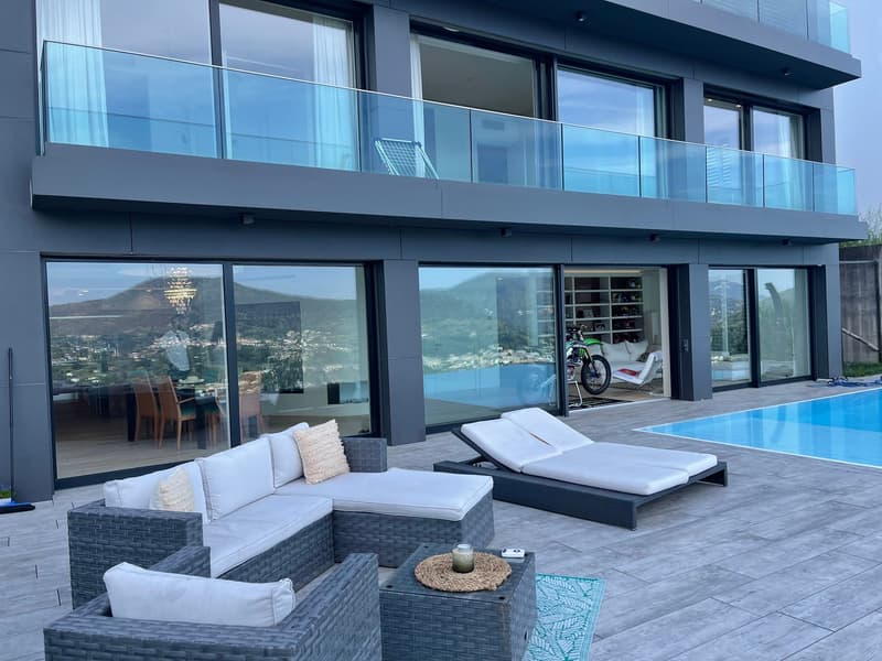 Modern two-story house with large glass windows and sliding doors, balcony, outdoor seating area with wicker furniture, swimming pool, and scenic mountain view
