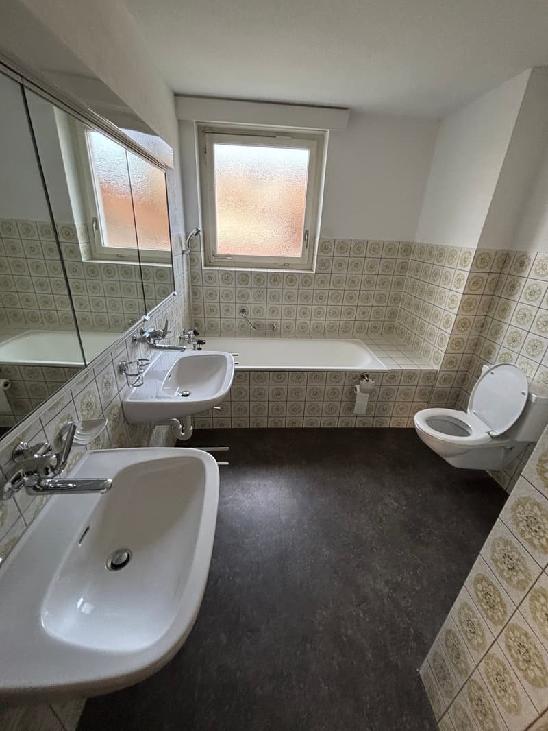 Tiled bathroom with white fixtures, including a bathtub, sink, and toilet. The floor is a dark colored tile. There is a window providing natural light.