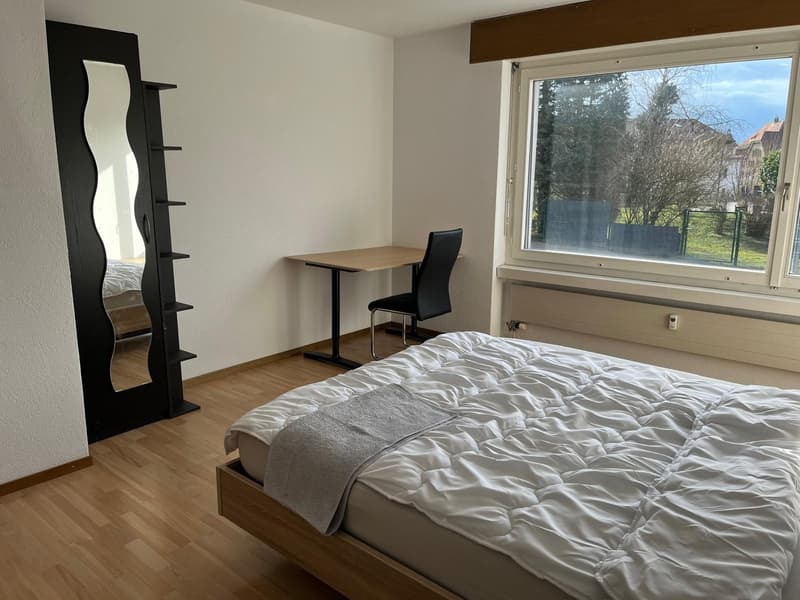 Spacious bedroom with large window overlooking outdoor greenery, wooden floor, black shelving unit, desk and chair