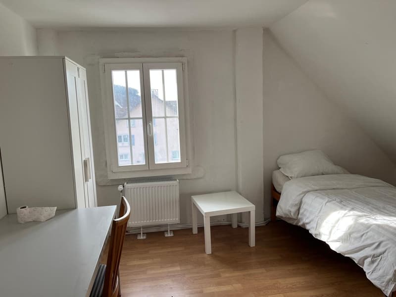 Bedroom with hardwood flooring, white walls, a double bed, a wardrobe, and a small table. There is a window providing natural light.