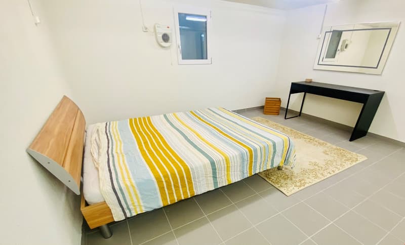bed with striped bedding, small rug, wall-mounted mirror, desk with drawers, window, air conditioning unit