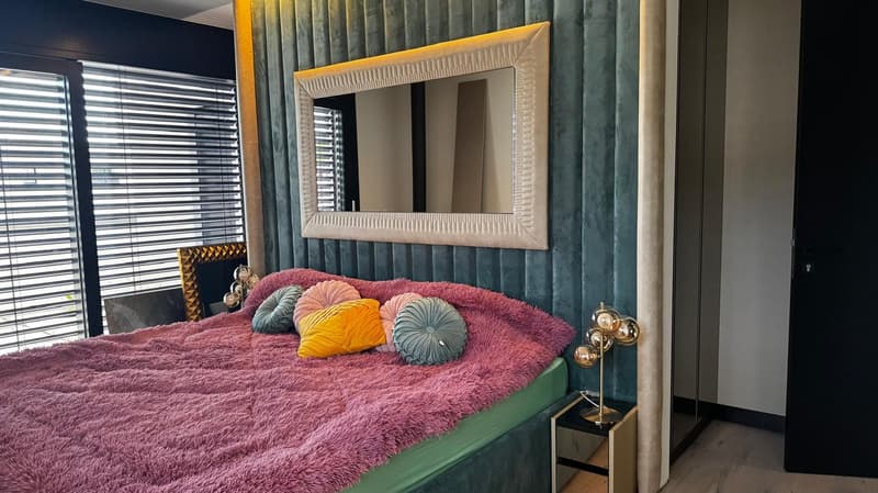 a bed with colorful cushions, a nightstand, a gold lamp, a large mirror, and a wooden floor