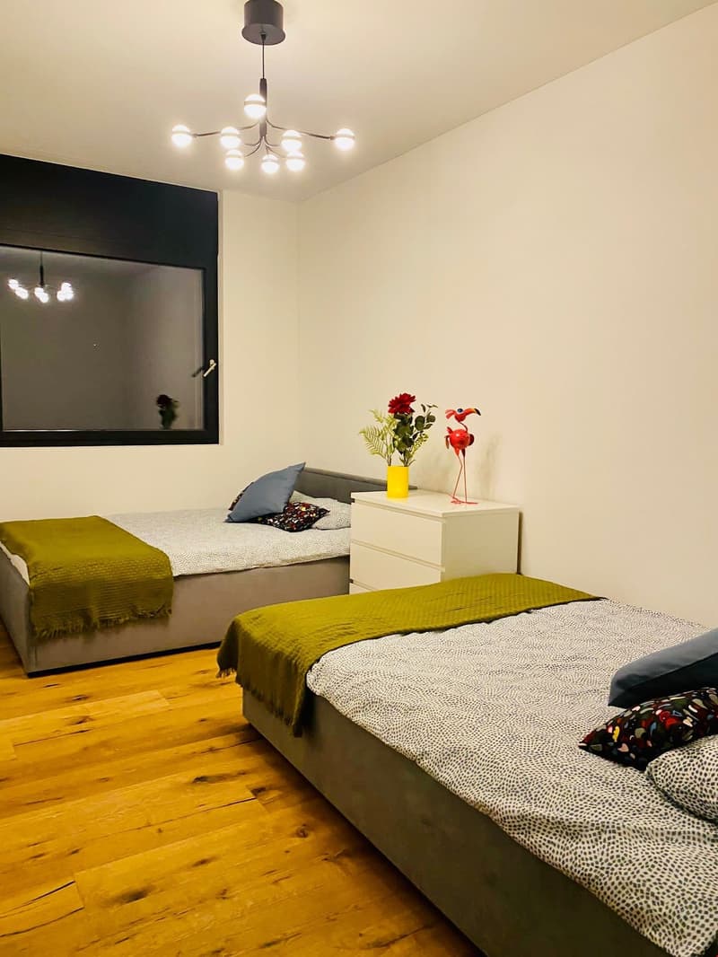 Two single beds, green blankets, white walls, a window with a reflection, a wooden floor, a decorative item on a side table, and a chandelier.