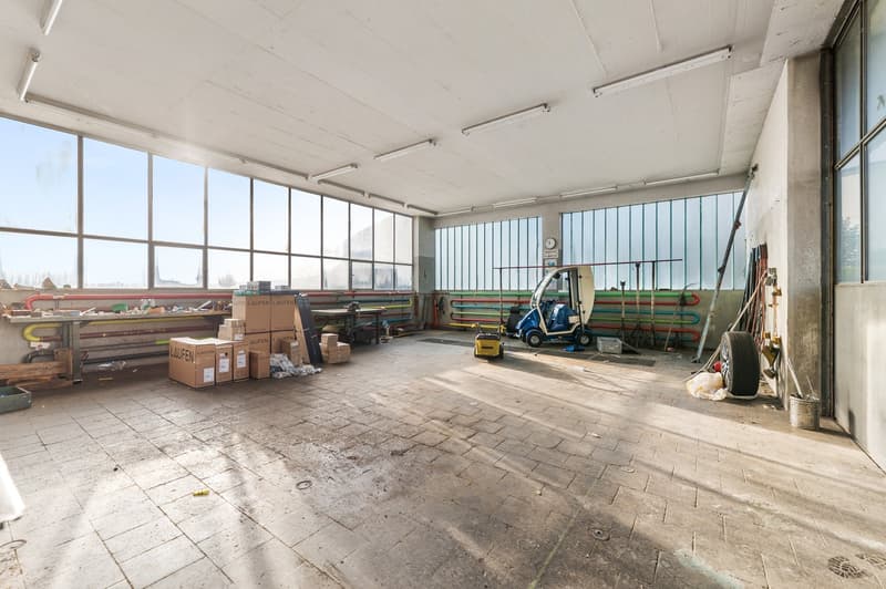Empty warehouse with large windows, concrete flooring, utility carts and boxes, ceiling lights