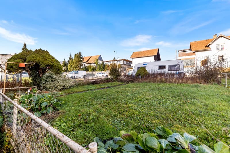 empty lot with fence, camper van, trees, shrubs, garden plot, road with cars in background