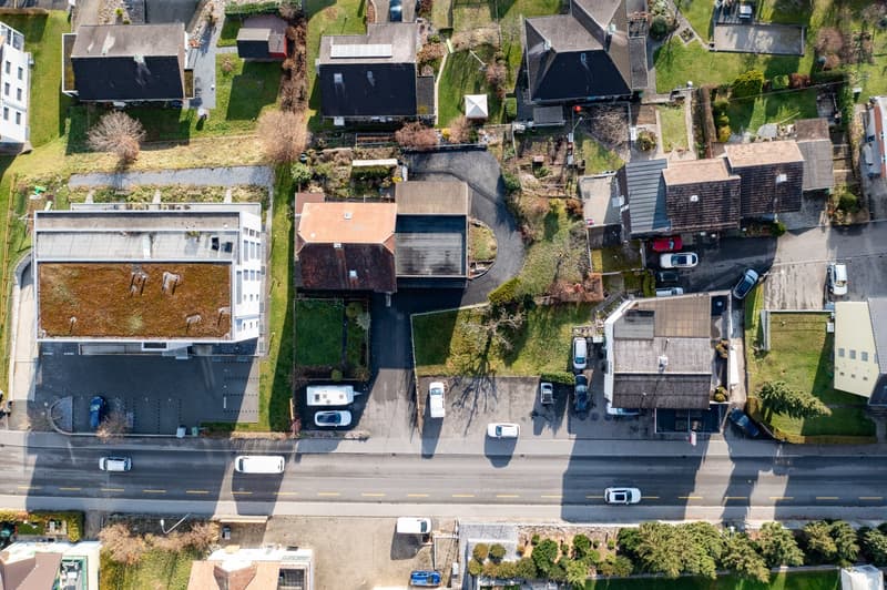 Aerial view of a residential area with multiple houses, driveways, cars parked, gardens, grassy fields, trees, and a street.