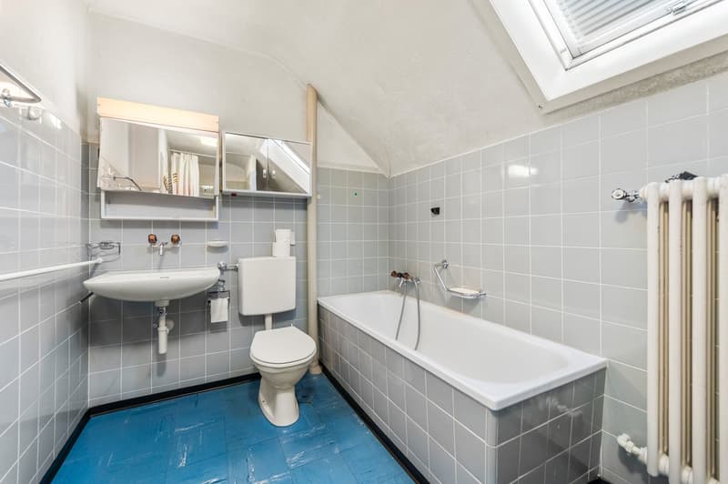 Bathroom with shower, toilet, and sink. White tiled walls and floor. A radiator is installed.