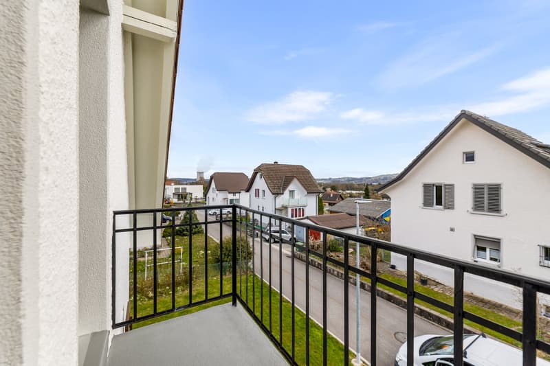 Balcony with black railings, overlooking a street with parked cars and houses