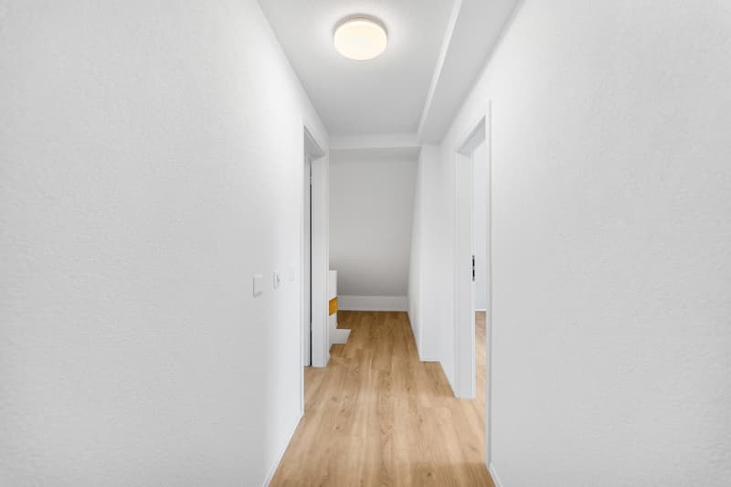 Empty hallway with white walls, wooden floor, and a ceiling light.