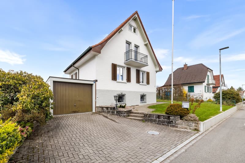 2 story house, white exterior, brown roof, garage, brick driveway, balcony, garden