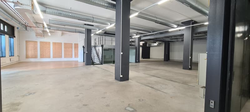 Large open commercial space with concrete flooring, exposed ceiling ducts, and support columns. The space appears to be unfinished, with plywood panels on the walls. There is a staircase visible in the background.