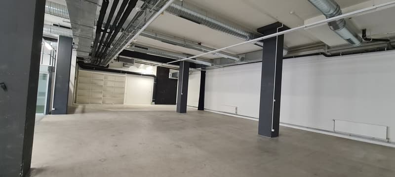 Large open commercial space with concrete flooring, exposed ceiling with ventilation ducts, and black support columns. The space appears to be unfinished or under renovation, with a clean and industrial aesthetic.